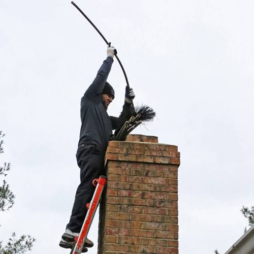 A person guiding a long chimney brush into a brick chimney from a ladder