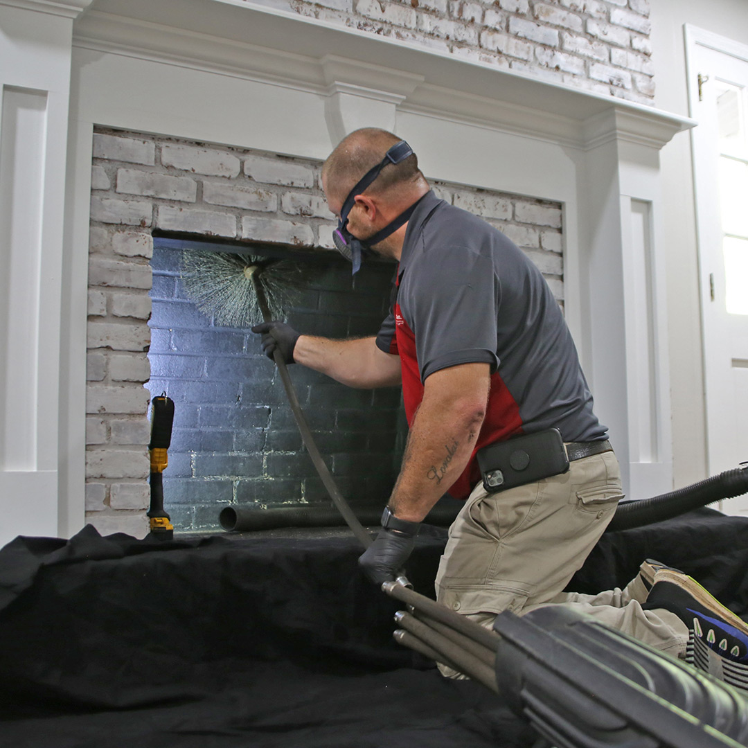 A person wearing a respirator cleans a white brick fireplace with a chimney brush